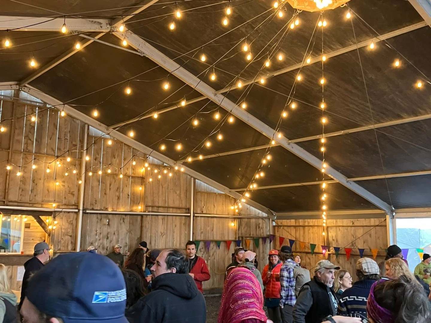 Crowd gathering under string lights in a rustic wooden barn with a pitched roof.