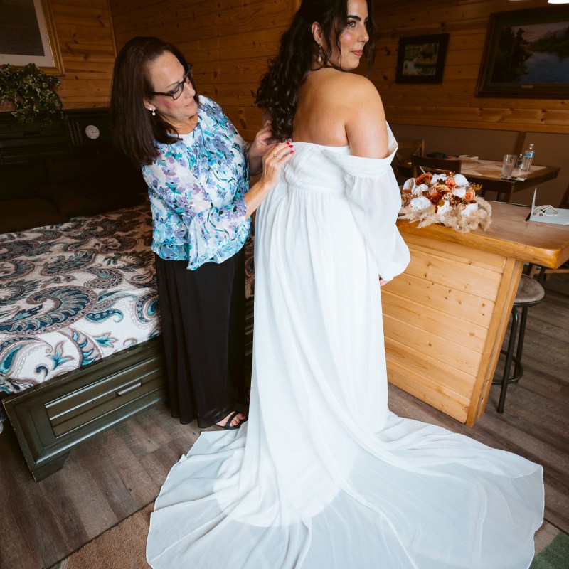 Woman helps bride fix her white dress in a wooden cabin interior.