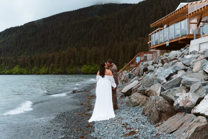 Couple in wedding attire kissing on a rocky beach by a lodge, with forested hills in the background.
