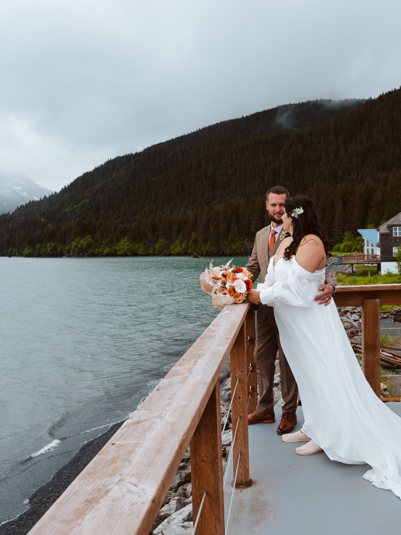 Couple on deck, bride in white gown, groom in brown suit, overlooking lake and forested mountains.