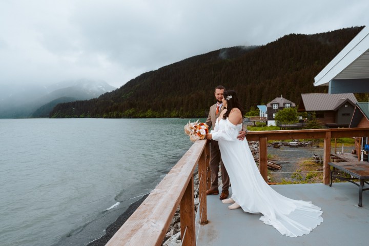 Couple on deck, bride in white gown, groom in brown suit, overlooking lake and forested mountains.