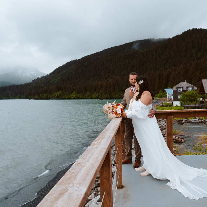 Couple on deck, bride in white gown, groom in brown suit, overlooking lake and forested mountains.