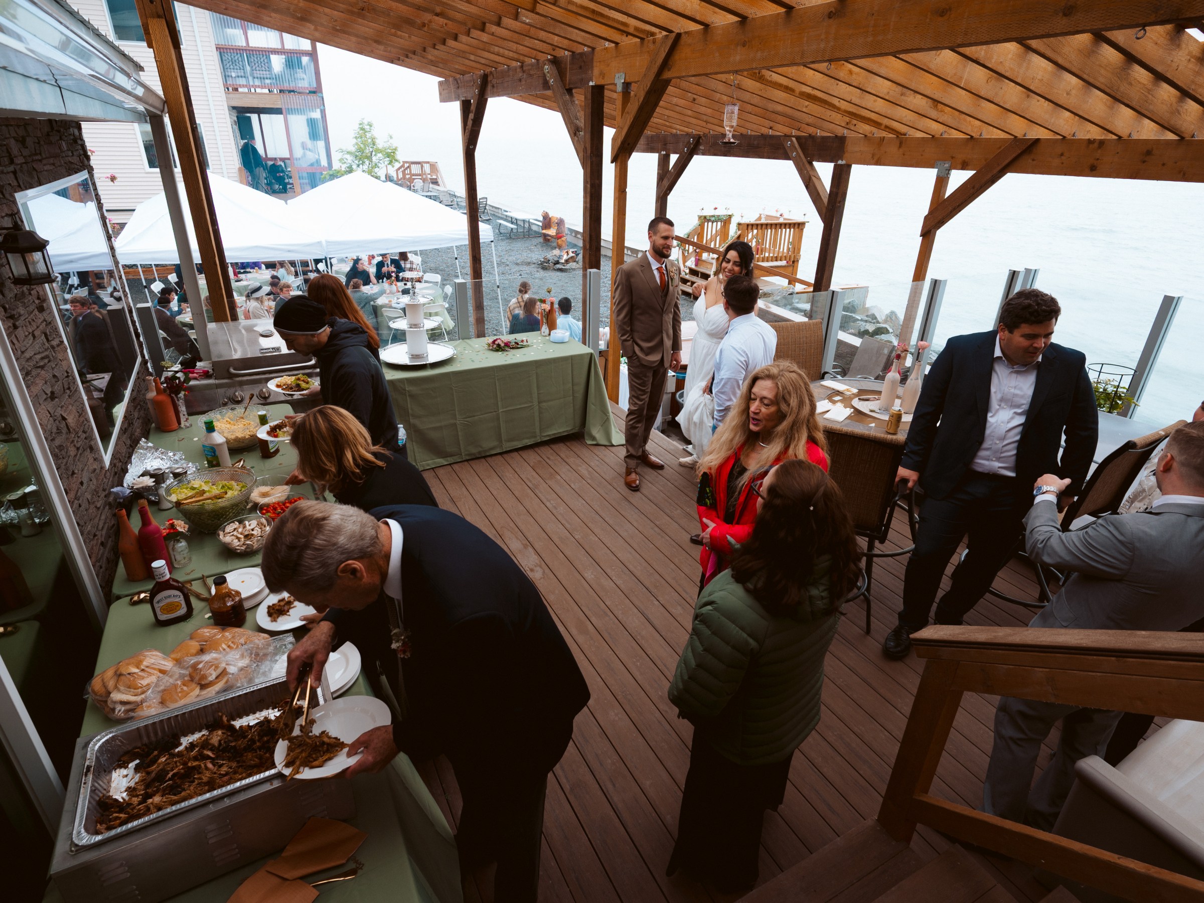 People at an outdoor buffet event on a wooden deck by the water.