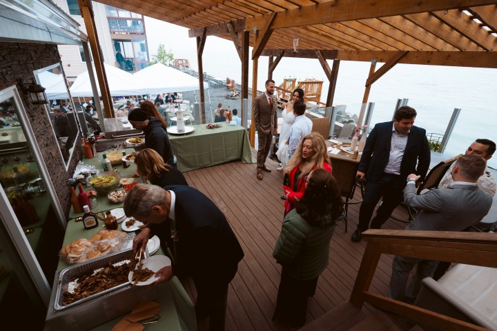 People at an outdoor buffet event on a wooden deck by the water.