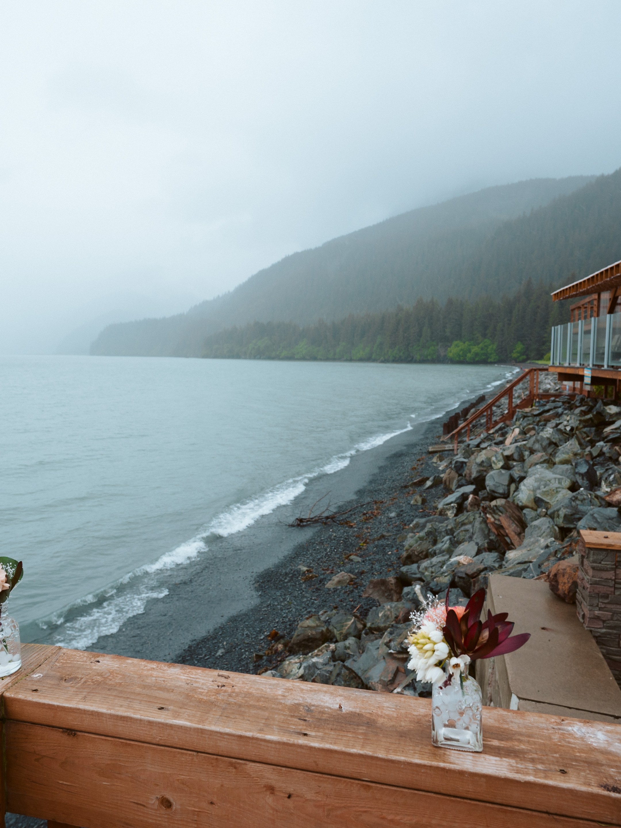 Rainy coastal view with wooden deck, flowers in vases, and forested mountains in the background.