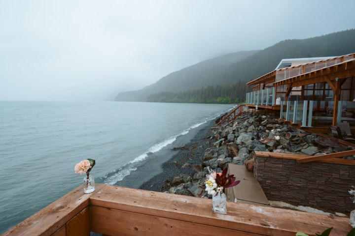Rainy coastal view with wooden deck, flowers in vases, and forested mountains in the background.