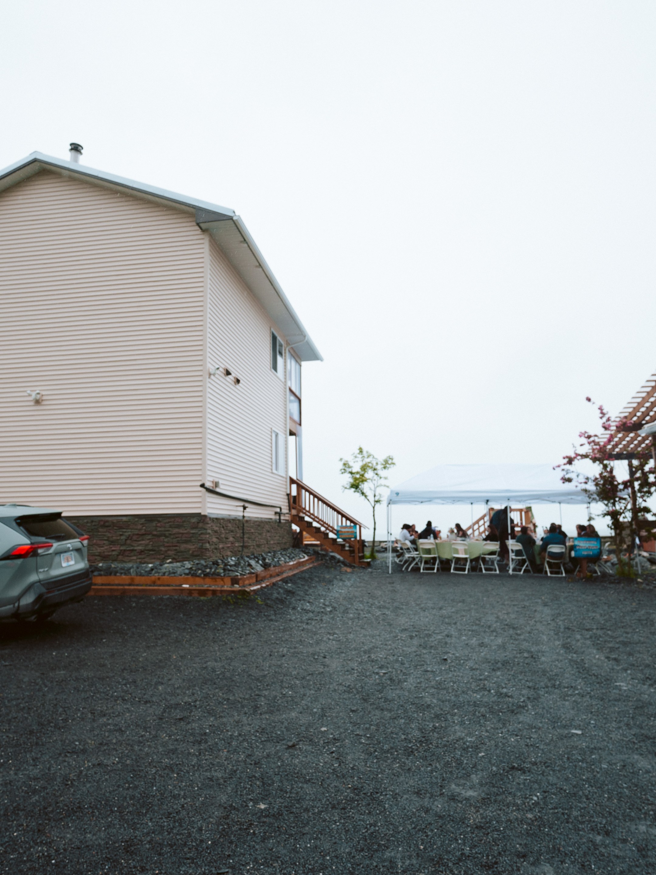 A house with parked cars, people under a white canopy, and wooden pergolas.