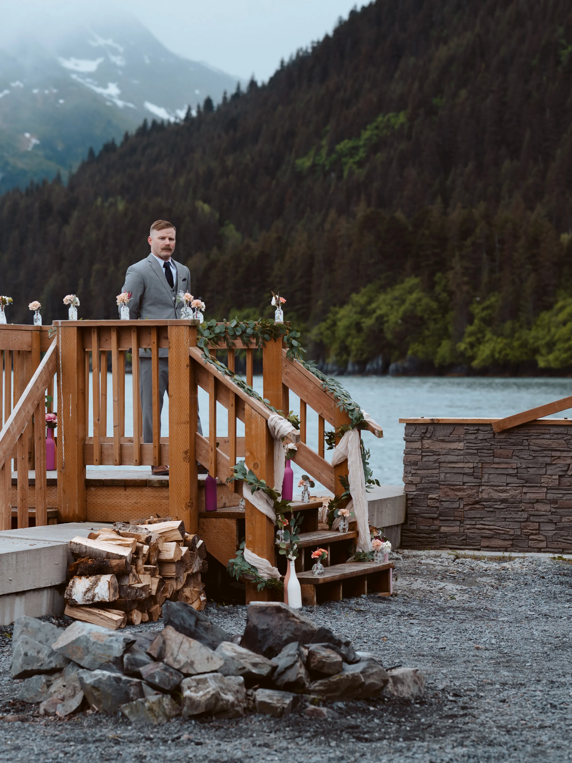 Man in suit on deck with floral decorations, overlooking a lake and forested mountain landscape.