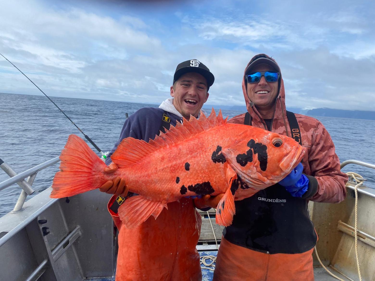 Two people on a boat holding a large orange fish with black spots, with ocean in the background.