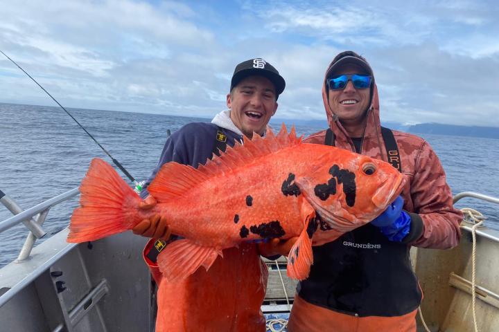 Two people on a boat holding a large orange fish with black spots, with ocean in the background.