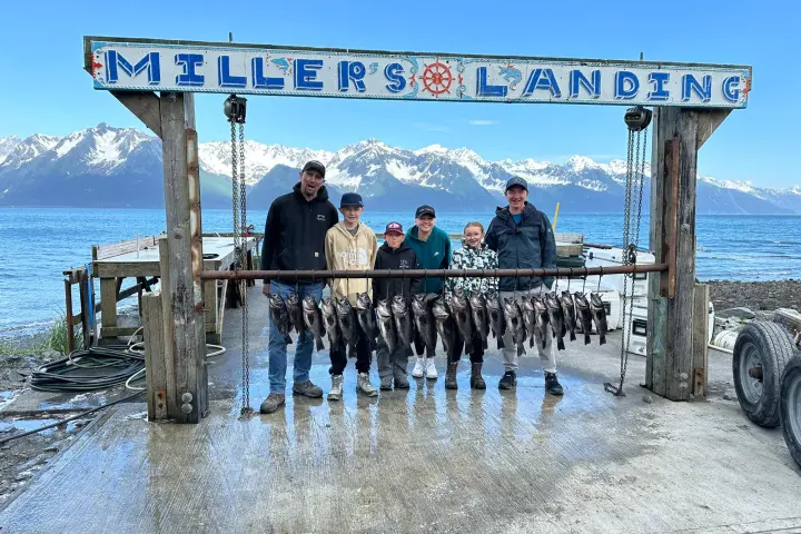 Group of people at Miller's Landing with caught fish hanging, snowy mountains and ocean in the background.