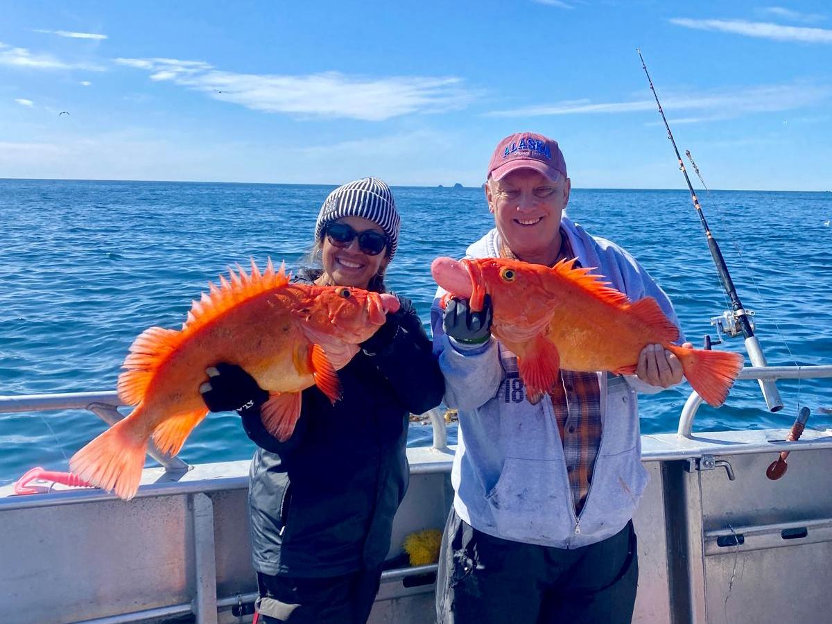 Two people on a boat holding large orange fish with the ocean in the background.