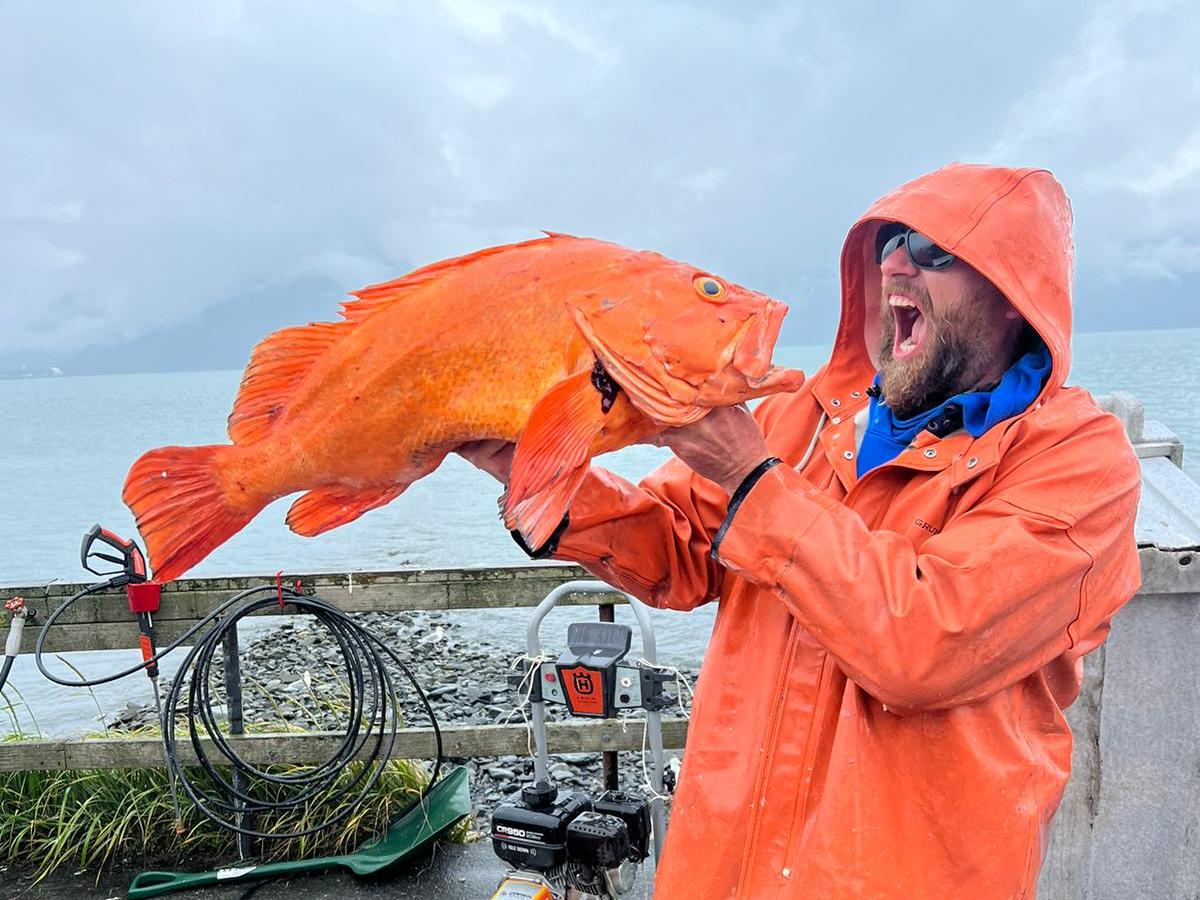 Person in orange rain gear holding a large orange fish by the coast.