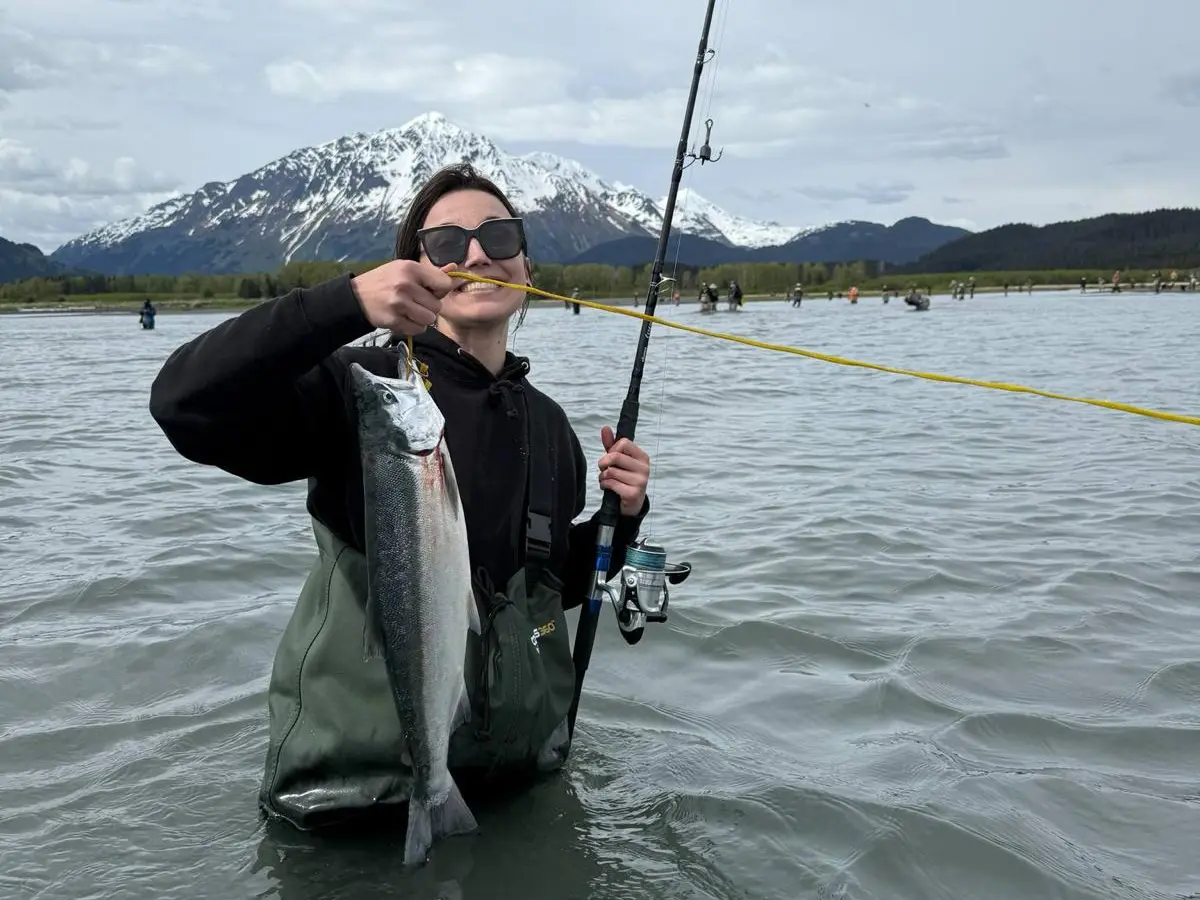 Person in water holding fish and fishing rod, with snowy mountain in the background.