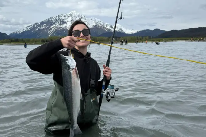 Person in water holding fish and fishing rod, with snowy mountain in the background.