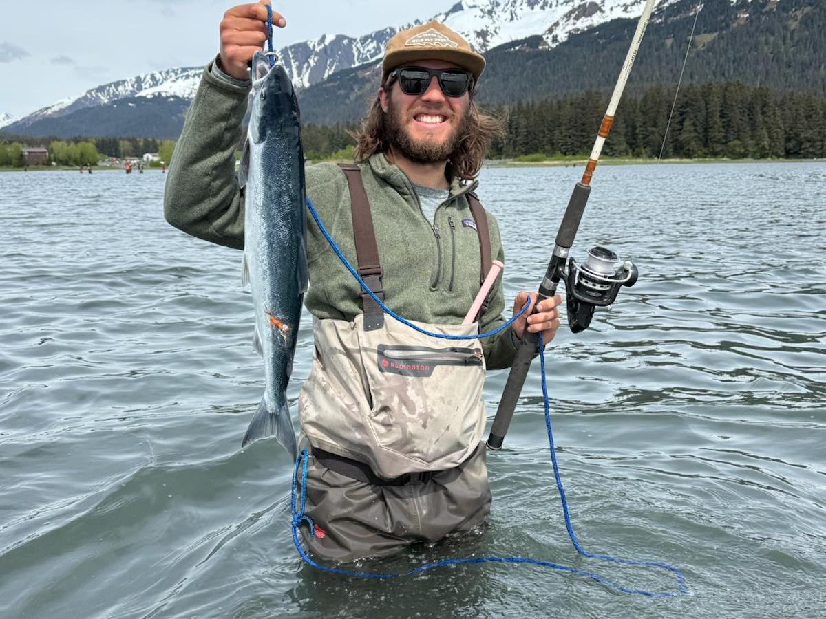Person in waders holding a fish and fishing rod, standing in water with snowy mountains in the background.