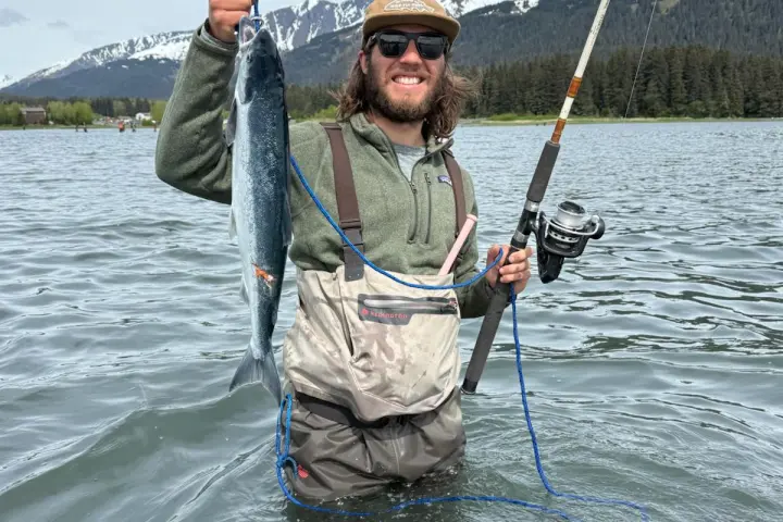 Person in waders holding a fish and fishing rod, standing in water with snowy mountains in the background.