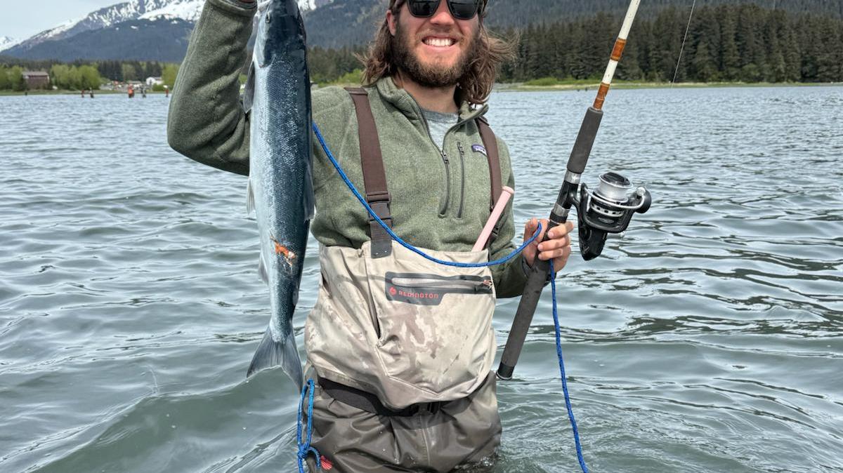 Person in waders holding a fish and fishing rod, standing in water with snowy mountains in the background.