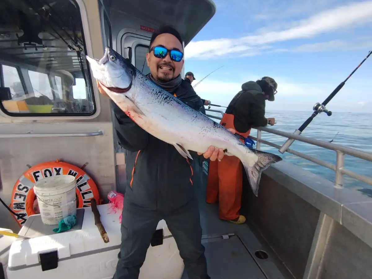 Smiling person on boat holding a large fish, with fishing rods and sea in the background.