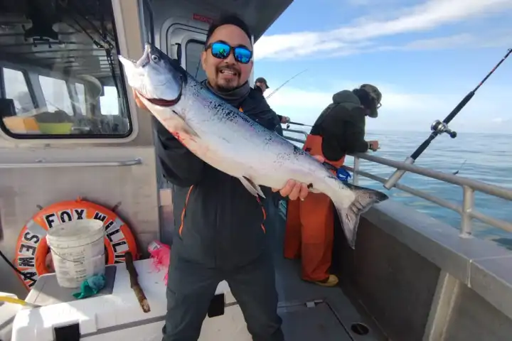 Smiling person on boat holding a large fish, with fishing rods and sea in the background.