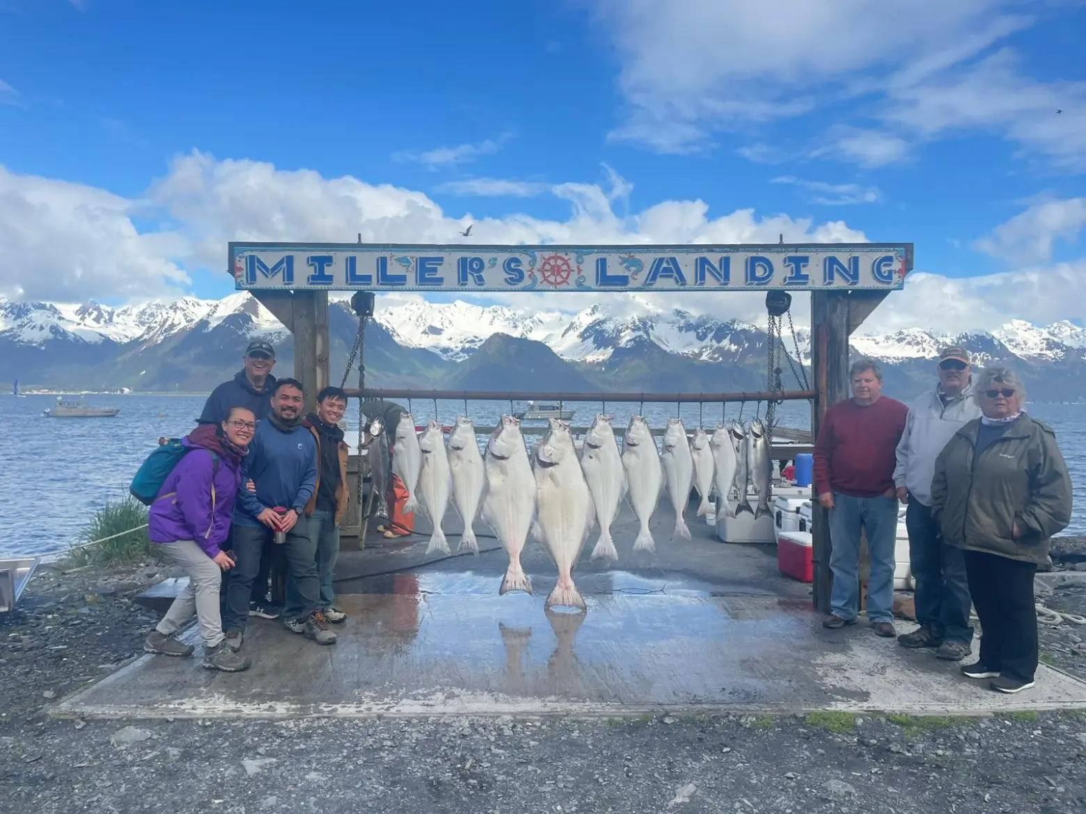 Group of people posing with caught fish under a sign that reads 'Miller's Landing' by a scenic mountain lake.