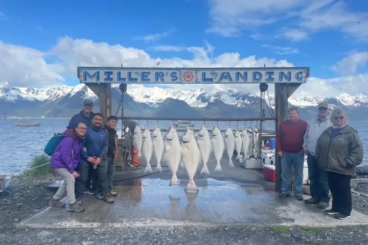 Group of people posing with caught fish under a sign that reads 'Miller's Landing' by a scenic mountain lake.