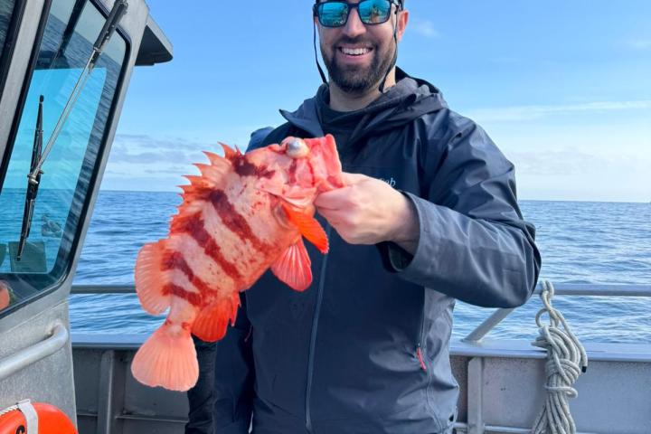 Person on a boat holding a red fish, wearing sunglasses and a cap, with a clear blue sky in the background.