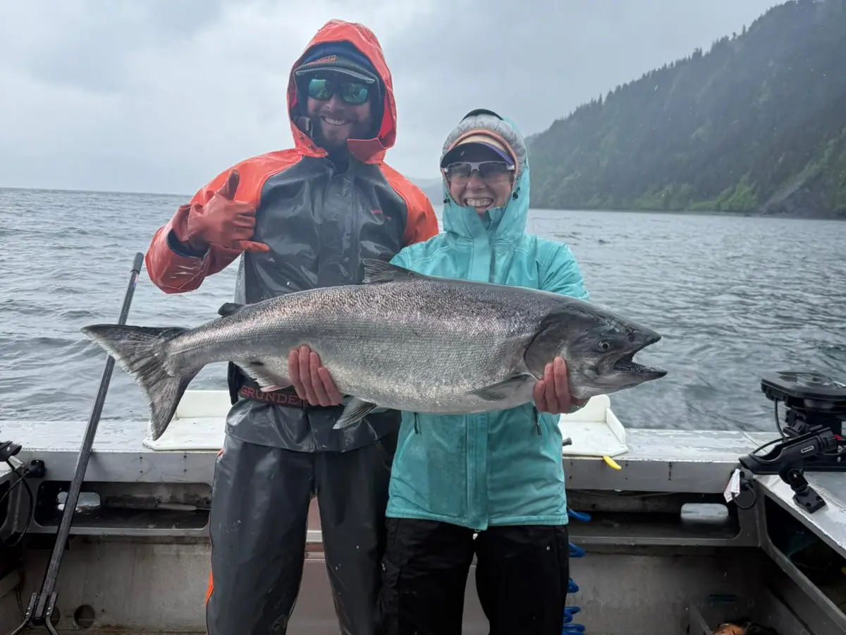 Two people in rain gear holding a large fish on a boat with a cloudy sky in the background.