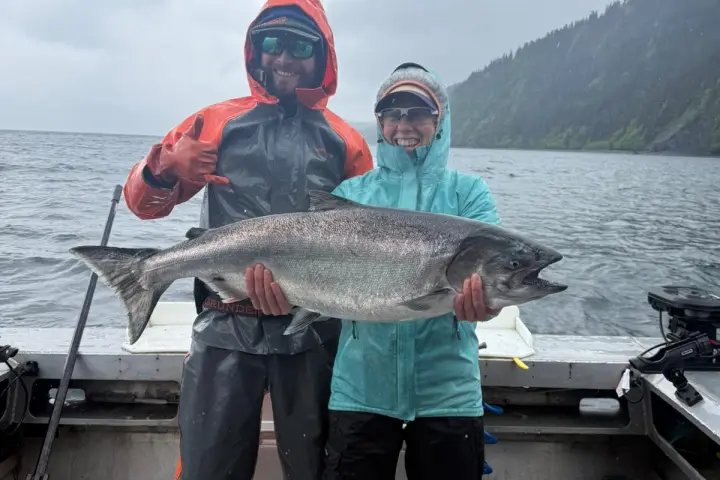 Two people in rain gear holding a large fish on a boat with a cloudy sky in the background.