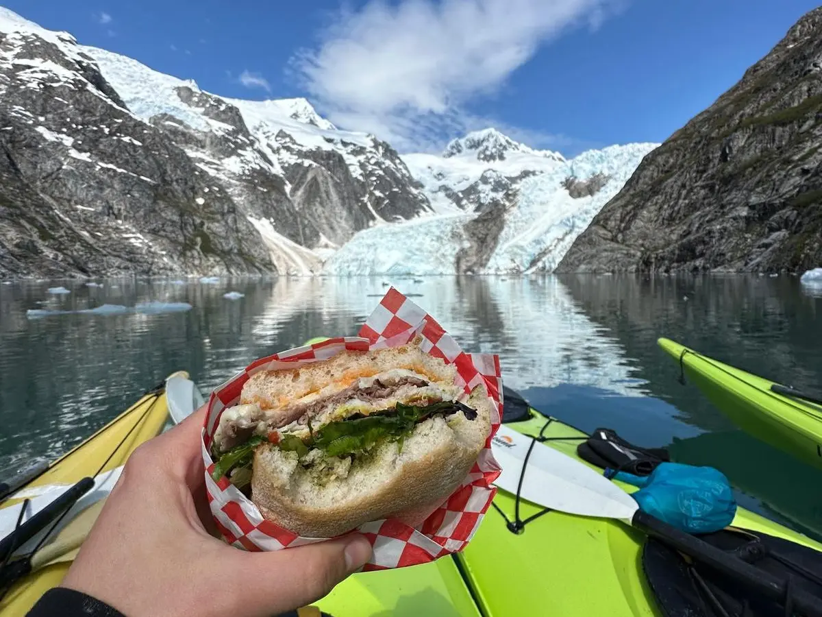 Person holding sandwich on kayak, with snowy mountains and bright sky in background.