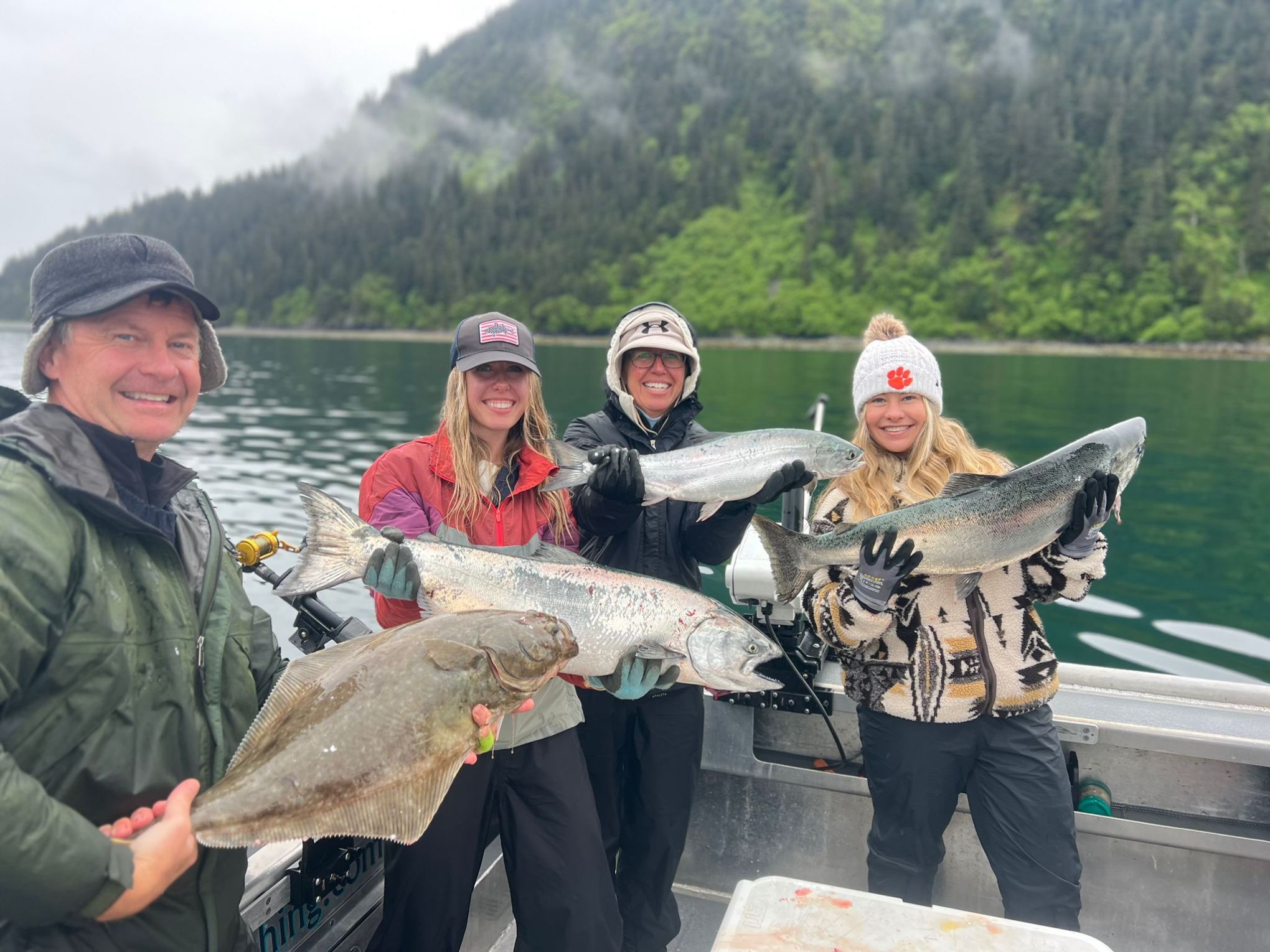 Four people on a boat holding large fish with a forested mountain backdrop.