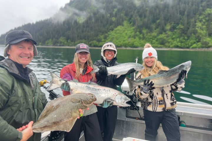 Four people on a boat holding large fish with a forested mountain backdrop.