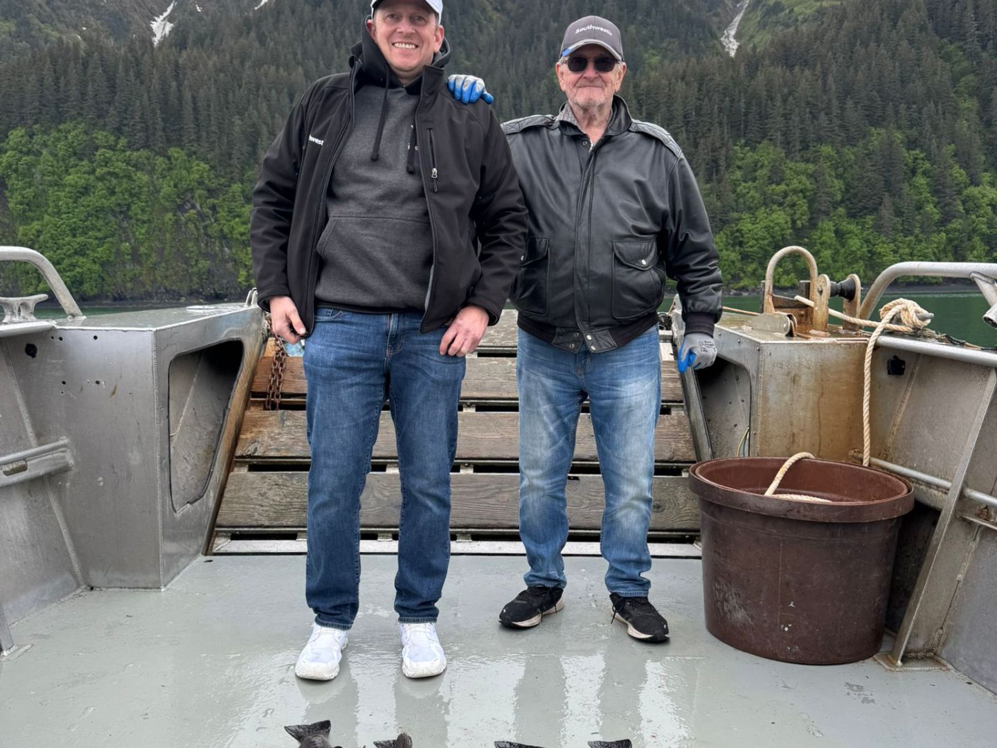 Two men standing on a boat deck with six large fish laid out, snowy mountains in the background.