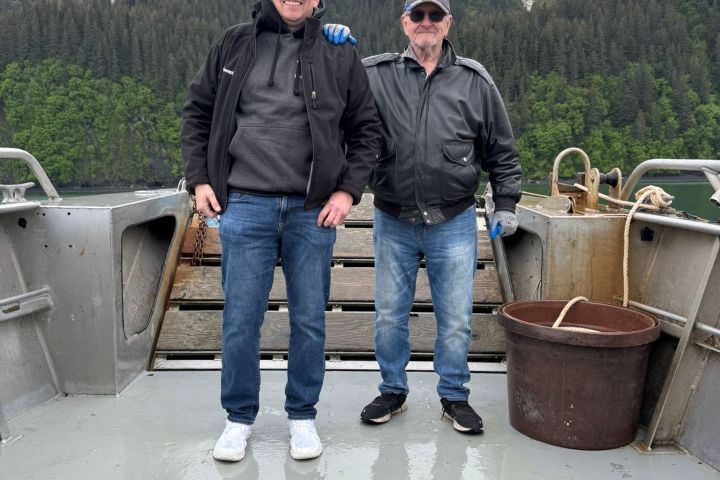 Two men standing on a boat deck with six large fish laid out, snowy mountains in the background.