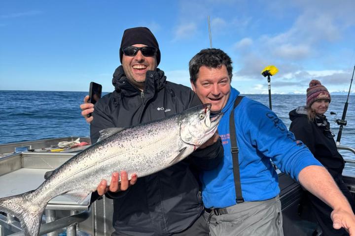 Two men on a boat holding a large fish, with the ocean and blue sky in the background.