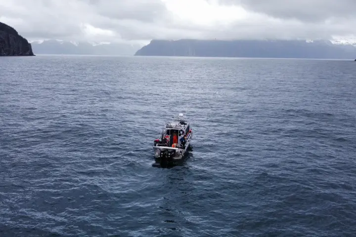 Boat on a calm sea with mountainous coastline under cloudy sky.