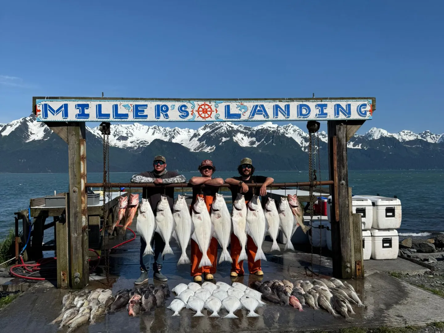 Three people display large fish at Miller's Landing with mountains in the background.