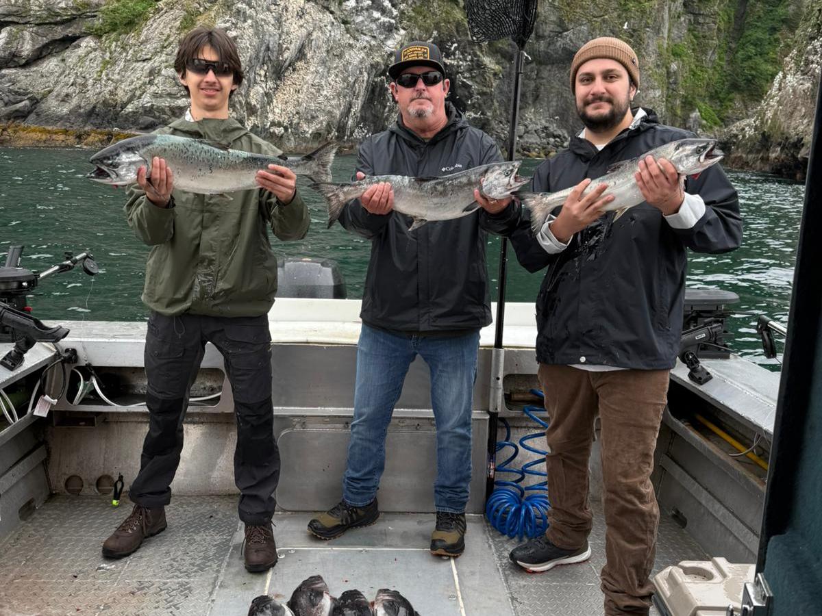 Three people holding fish on a boat, with more fish on the boat floor and cliffs in the background.