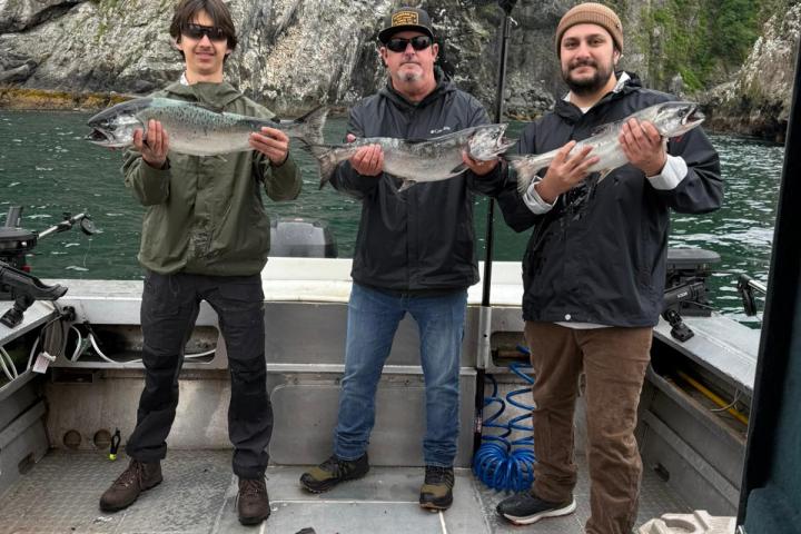 Three people holding fish on a boat, with more fish on the boat floor and cliffs in the background.