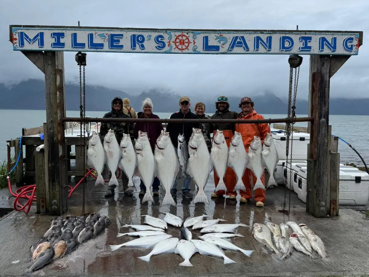 Group stands behind large hanging fish at Miller's Landing fishing site, overcast sky in background.
