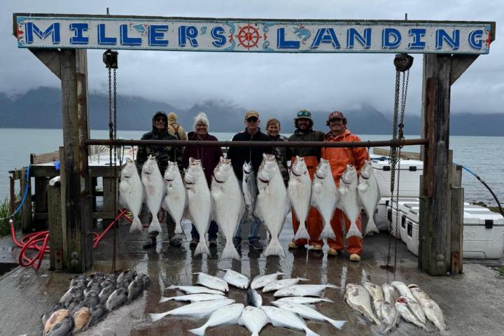 Group stands behind large hanging fish at Miller's Landing fishing site, overcast sky in background.