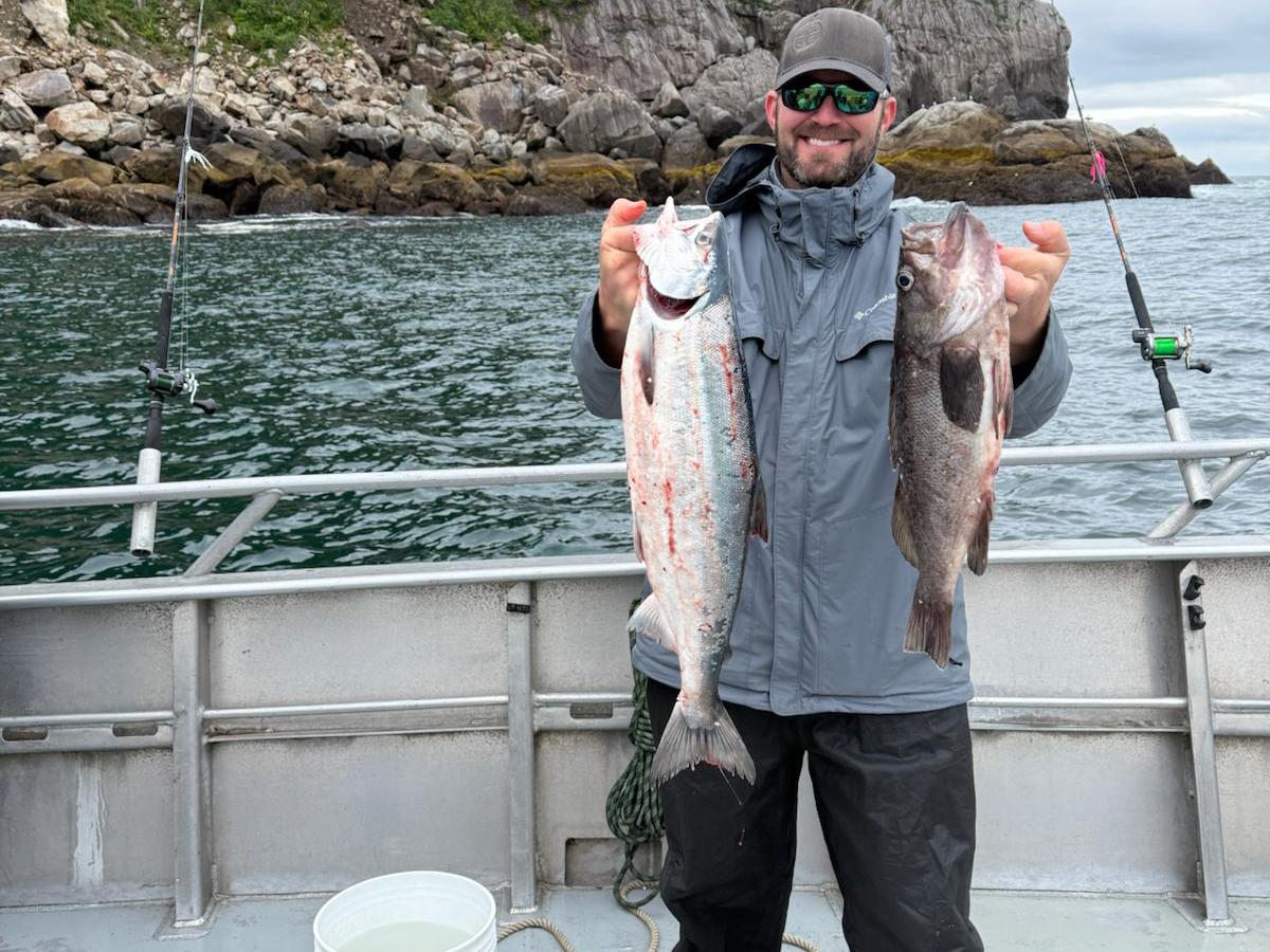 Person on a boat holding two fish, with rocky cliffs and trees in the background.