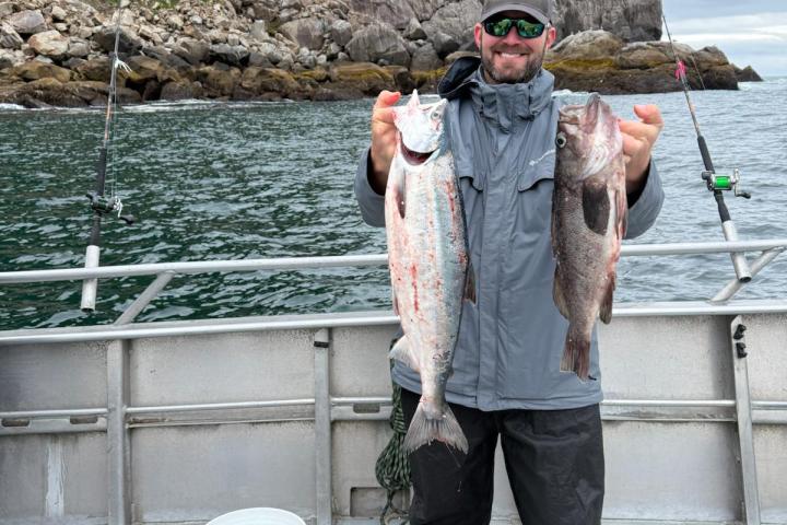 Person on a boat holding two fish, with rocky cliffs and trees in the background.