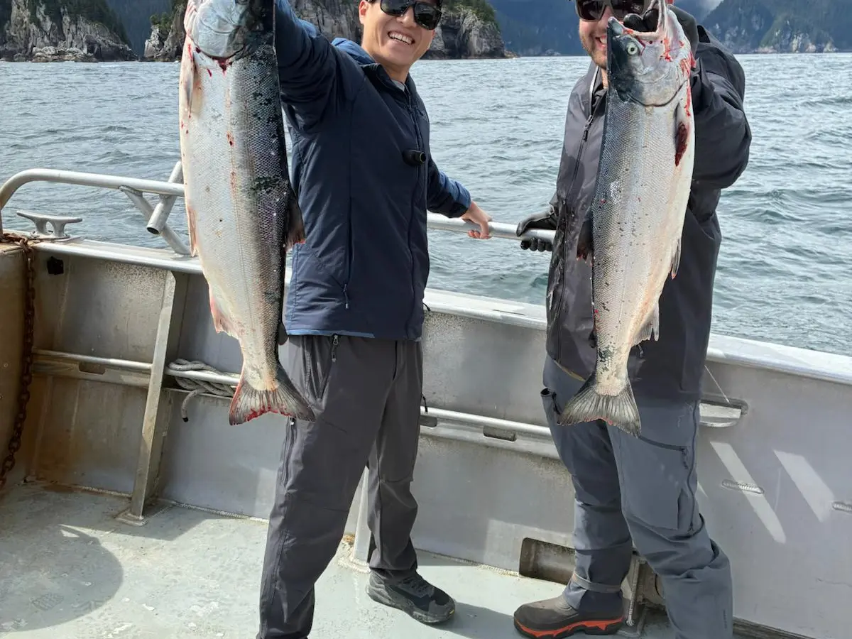 Two people on a boat holding large fish with a scenic coastline in the background.