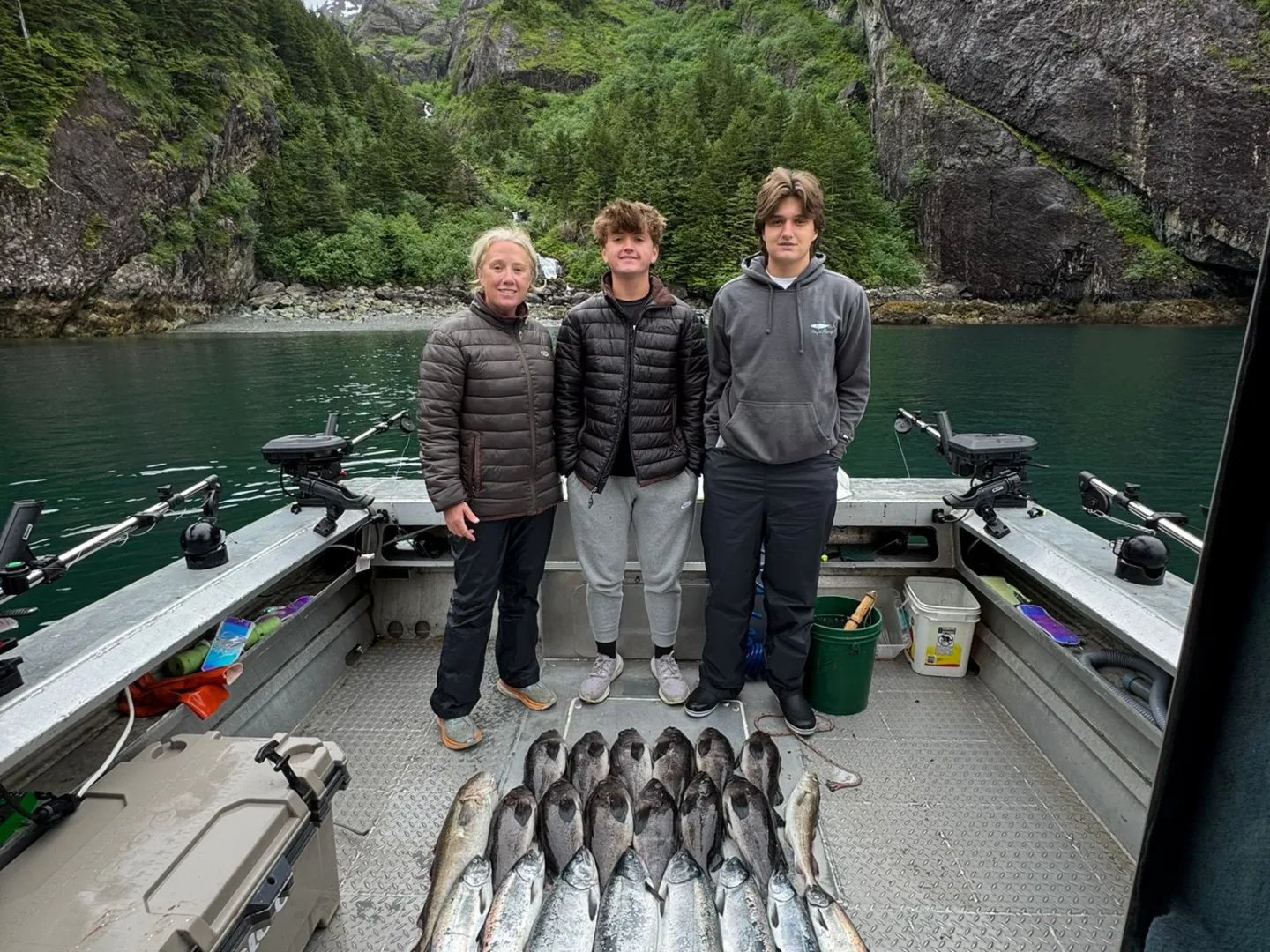 Three people stand on a boat with a large catch of fish, surrounded by tall cliffs and forested scenery.