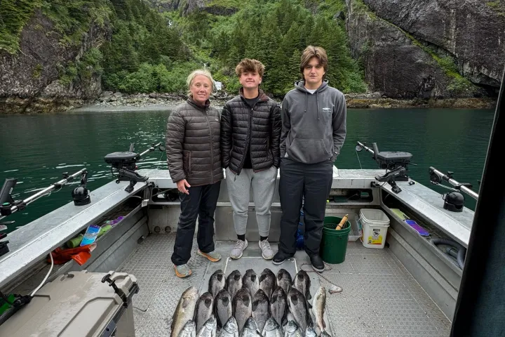 Three people stand on a boat with a large catch of fish, surrounded by tall cliffs and forested scenery.