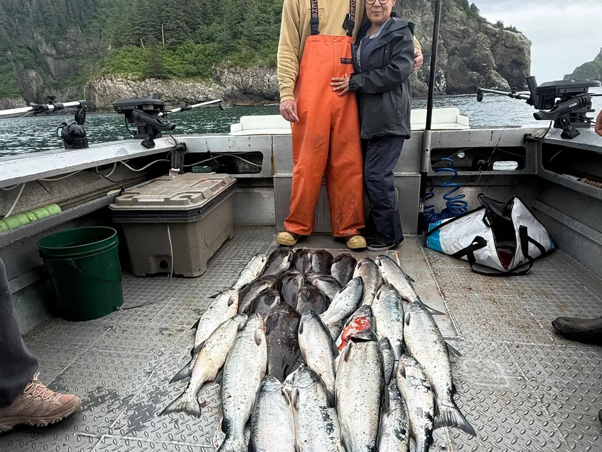Two people posing on a boat with a large catch of fish, forested cliffs in the background.