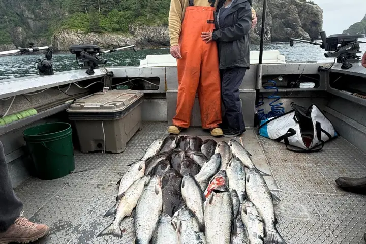 Two people posing on a boat with a large catch of fish, forested cliffs in the background.