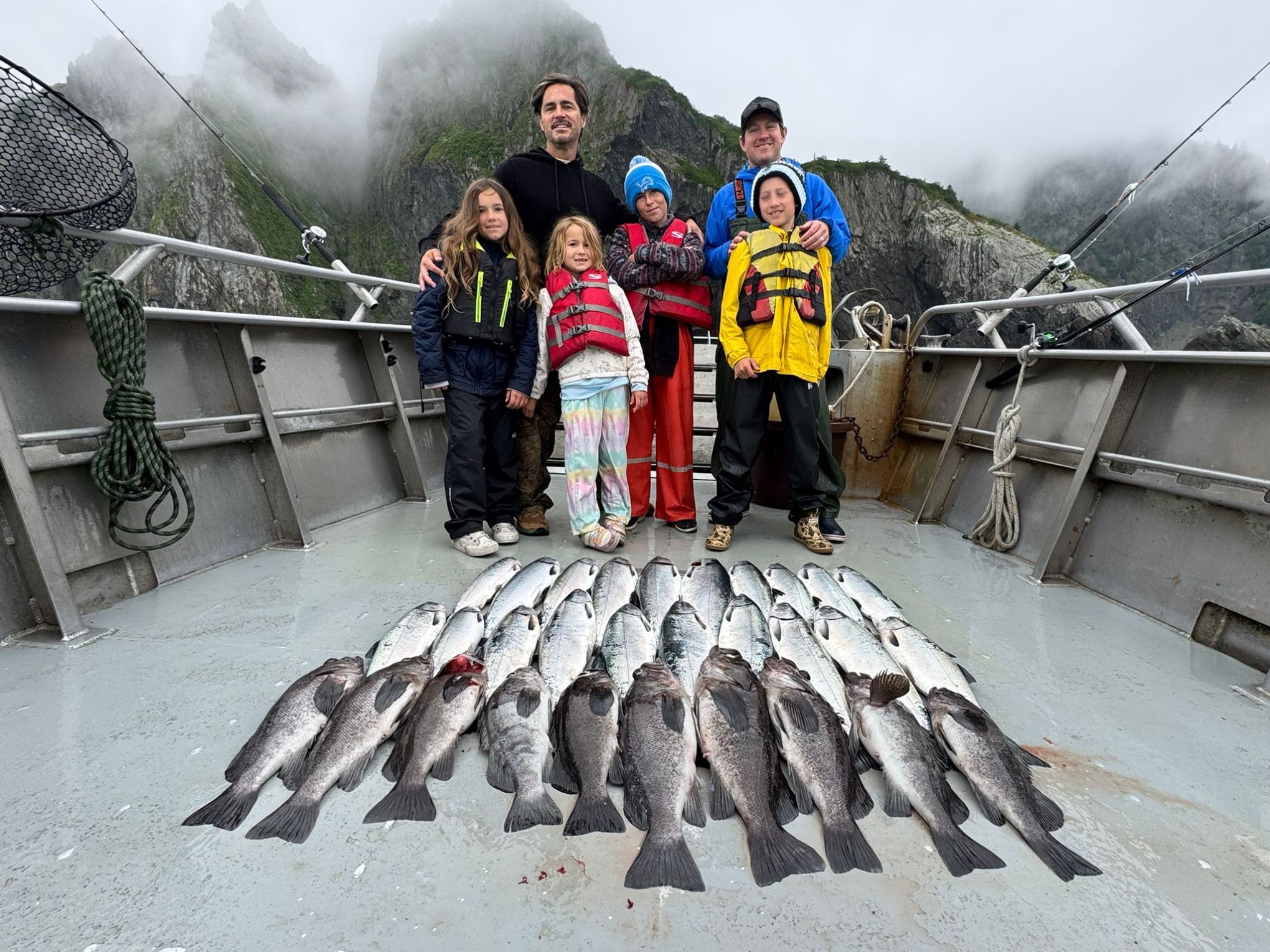 Group on boat with fishing gear and many fish, mountains in background.