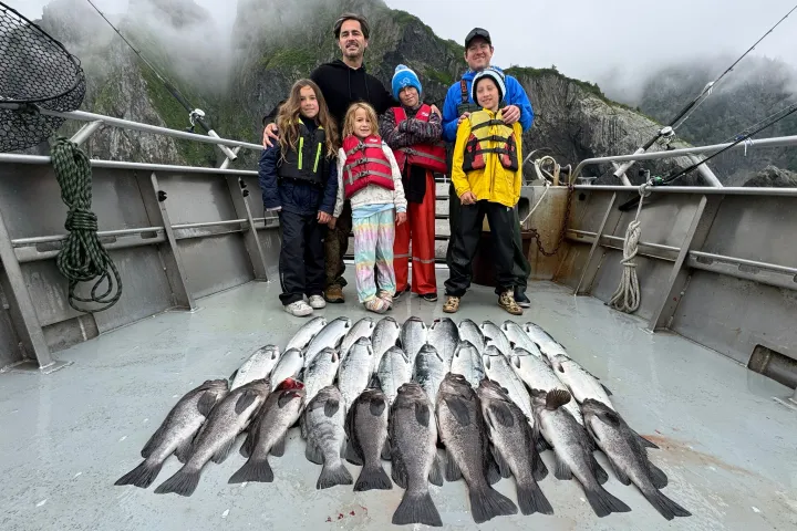 Group on boat with fishing gear and many fish, mountains in background.